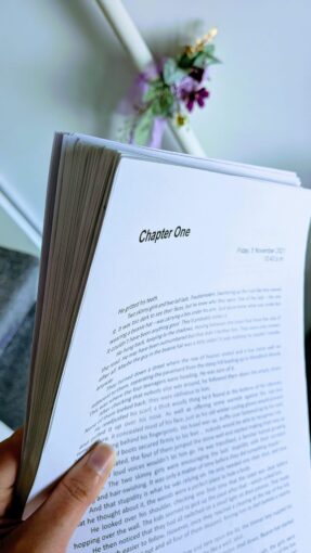 A close-up shot of a pair of hands holding a well-read book manuscript, with a purple heart and pen emoji in the background.